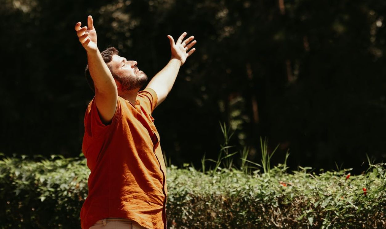 A man stands outside with arms raised, embracing the sunlight in a park setting