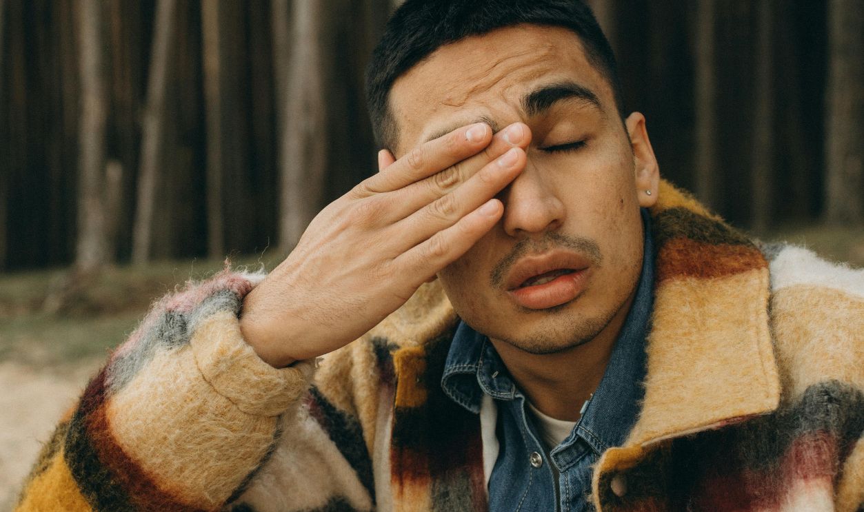 Young man in stylish jacket rubbing his eye in a forest setting, capturing a moment of introspection