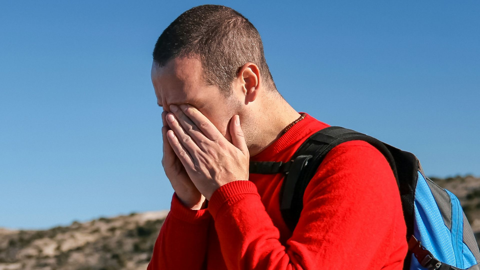 man wearing red crew-neck sweater with teal and black backpack outdoor during daytime