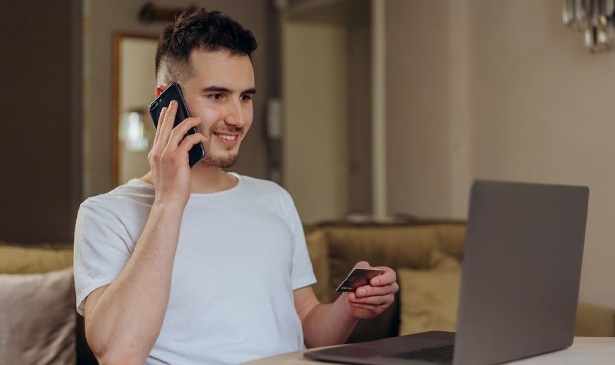 A young man smiles while shopping online using a phone and laptop, holding a credit card.