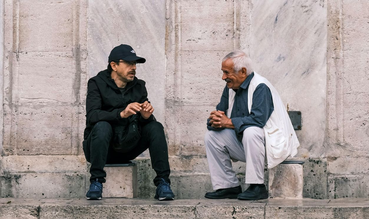 Two men sitting on stone steps engaging in conversation against a marble wall