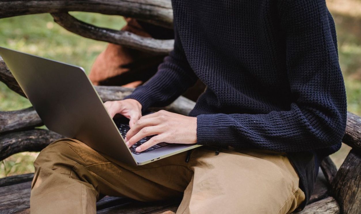A man sitting on a wooden bench using a laptop outdoors in a park during daytime.