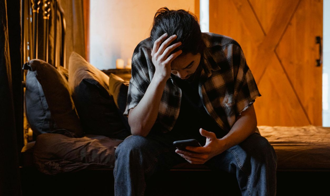 Young man in a flannel shirt sitting on bed, looking at phone, surrounded by bottles, in a dimly lit room
