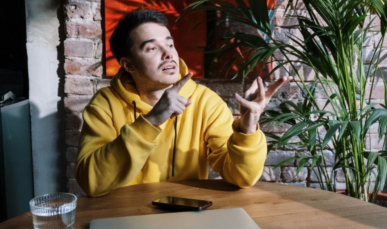 Young man in a yellow hoodie gesturing while sitting at a table with a laptop and cellphone