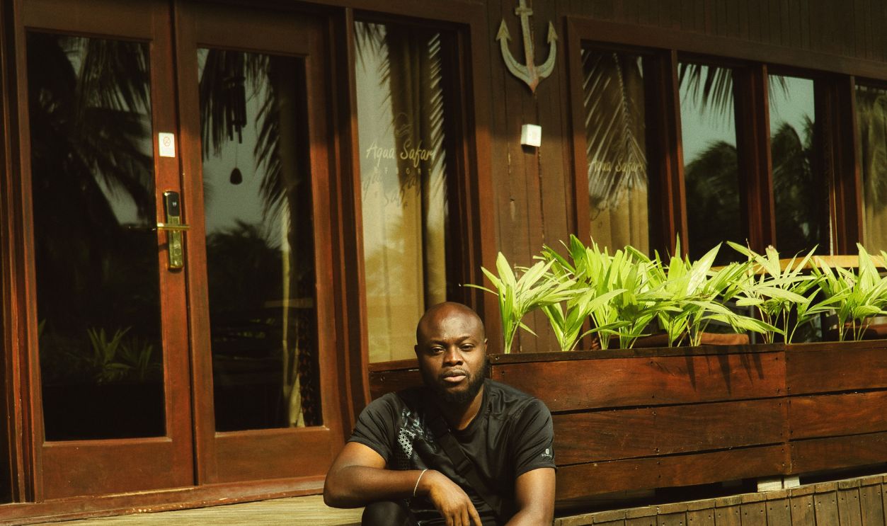 Man sitting on wooden cabin steps in Ada Foah, Ghana, with tropical plants