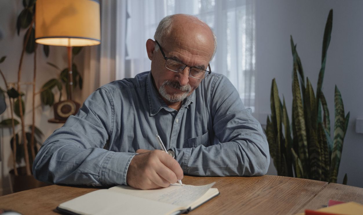 A senior adult man writing in a notebook at a desk in a cozy home office setting