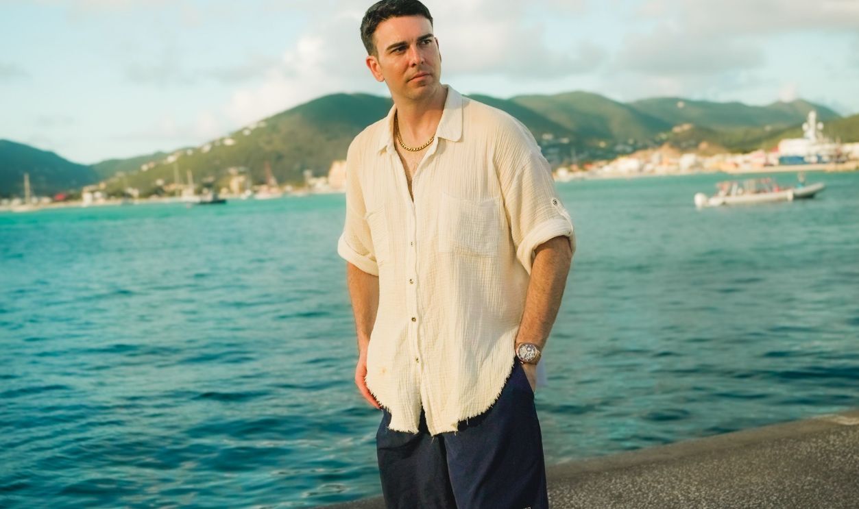 A man stands by the ocean with hills in the background, enjoying a sunny day