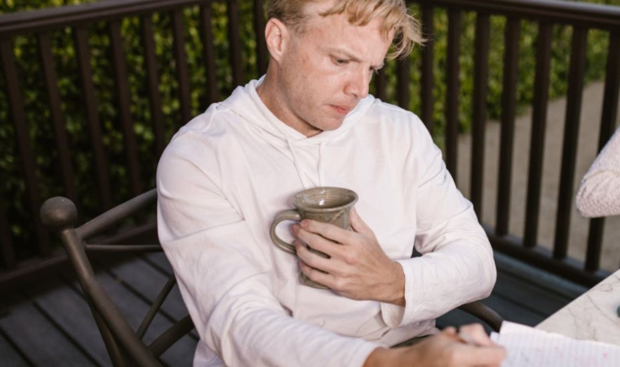 Man in a hoodie writing in a notebook while holding a mug, sitting outdoors