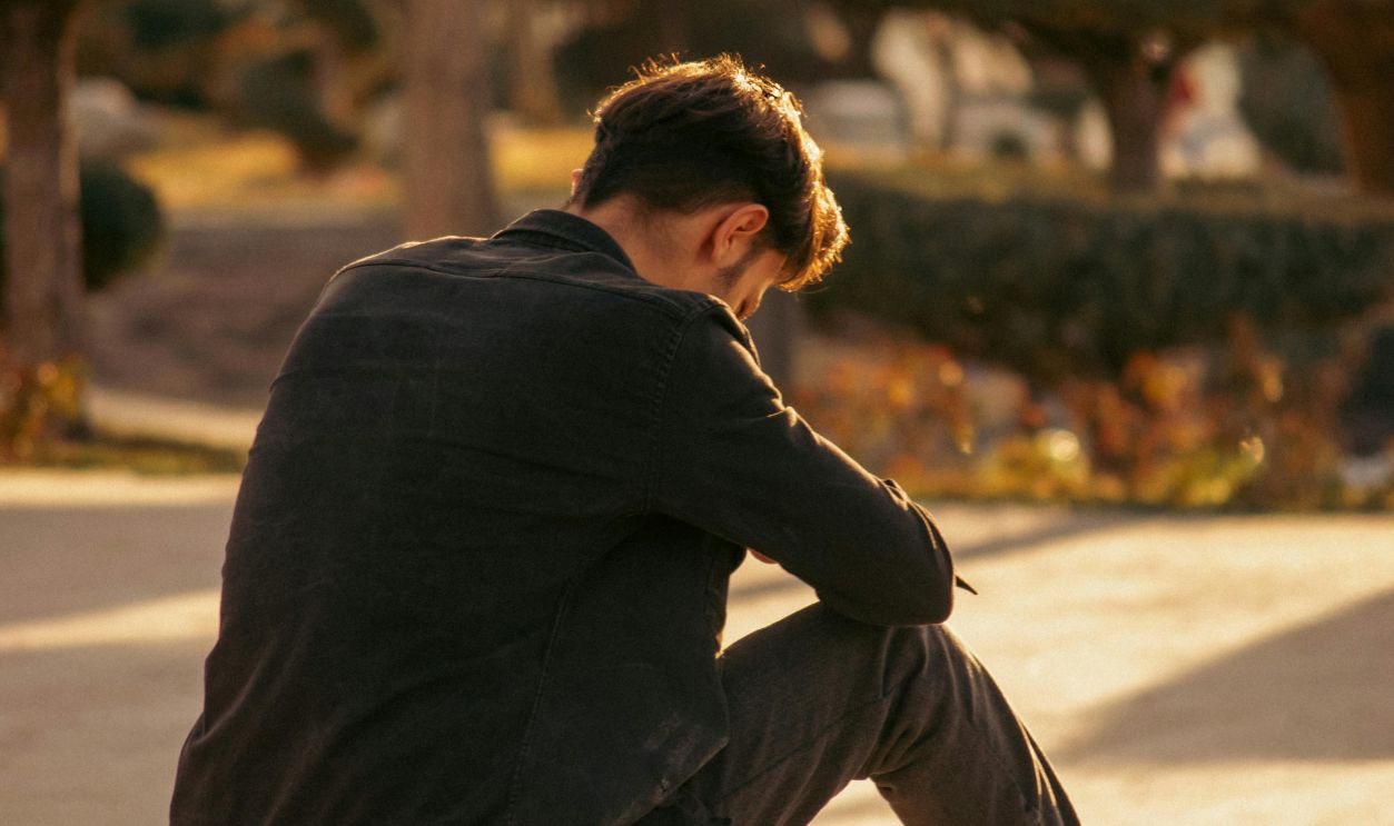 A young man sits alone in a park, deep in thought, during a sunny autumn day