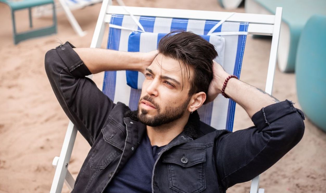 A bearded man in a denim jacket relaxing on a beach chair by the sand