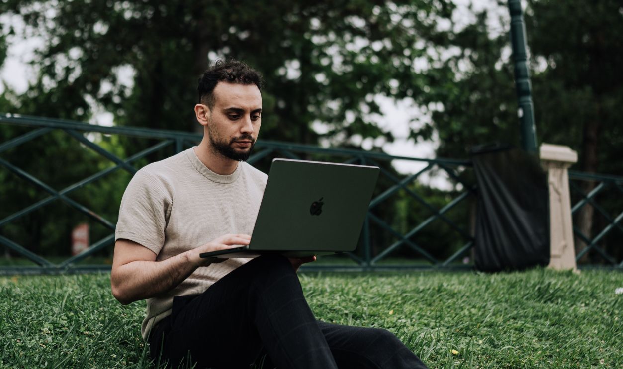 A young man sitting on grass using a laptop in a serene park setting, showcasing portable technology