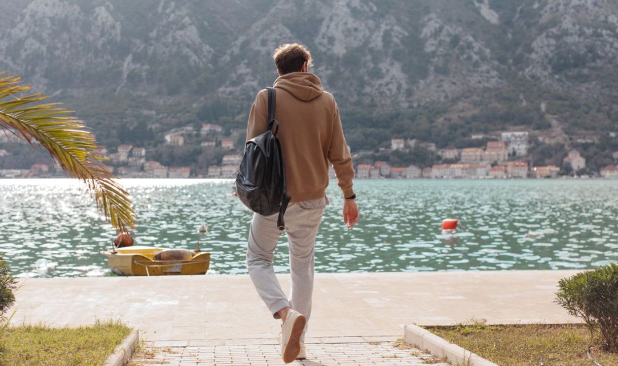 Man in a hoodie walking on a dock towards calm waterfront with mountains in the background