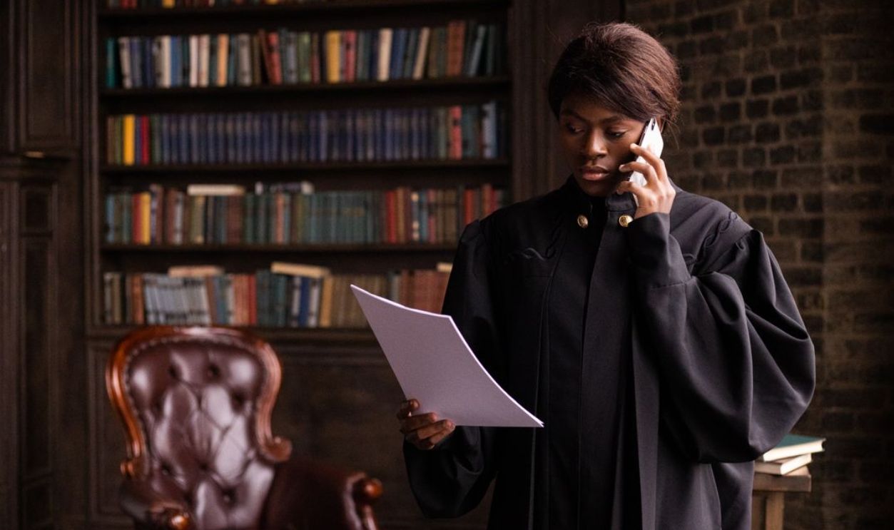 African American judge in traditional robes holds a document and talks on the phone in a law library