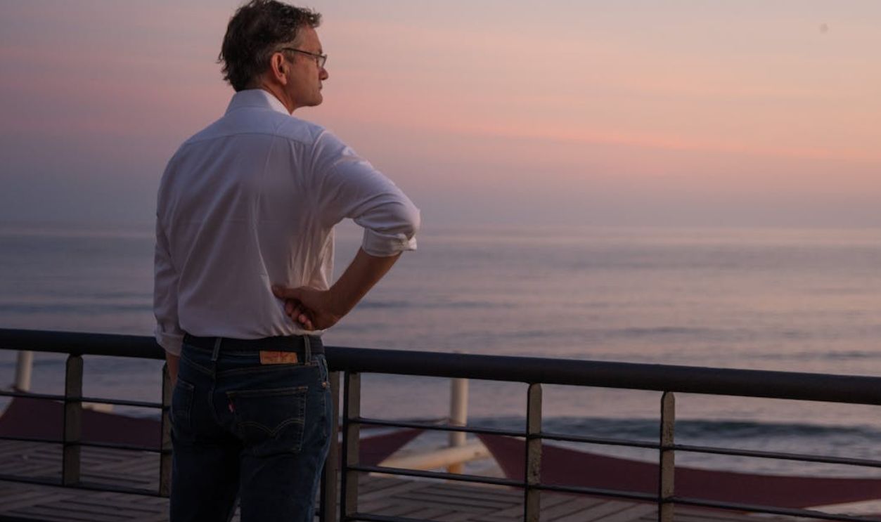A man in a white shirt gazes at the ocean during a peaceful sunset, exuding calmness.