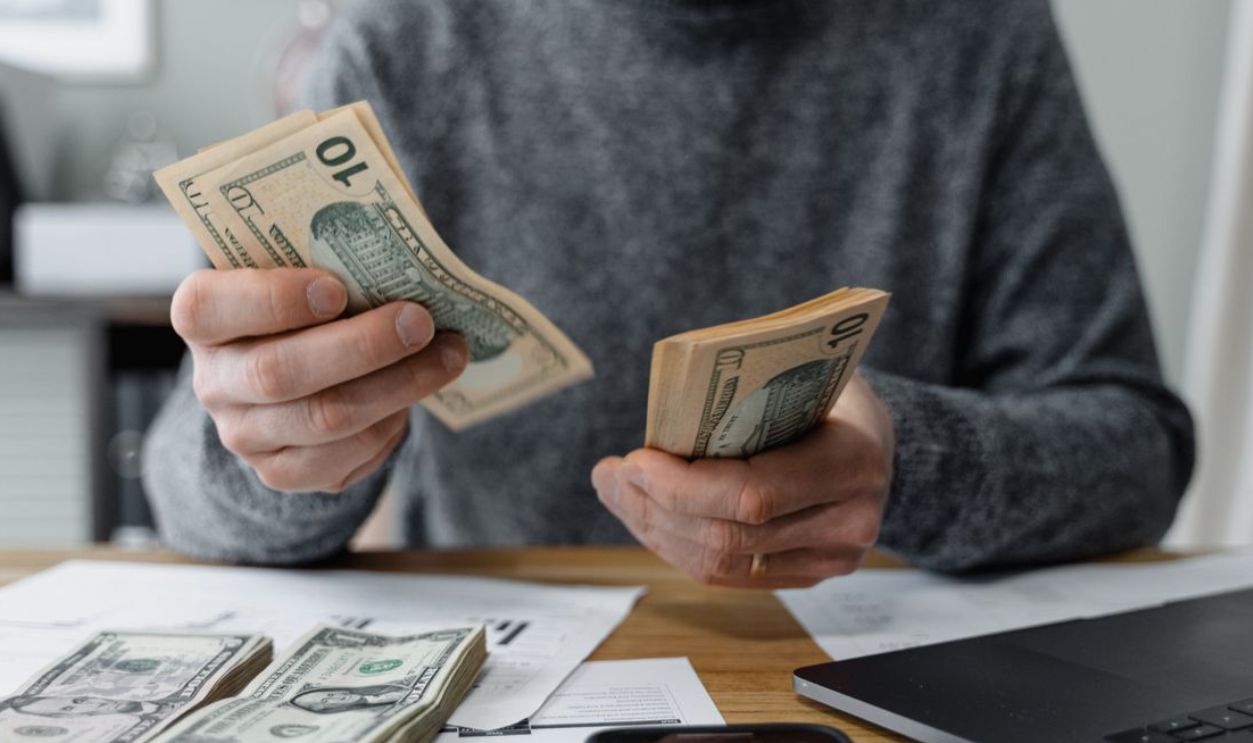 Close-up of person counting cash with a calculator and paperwork on a desk.