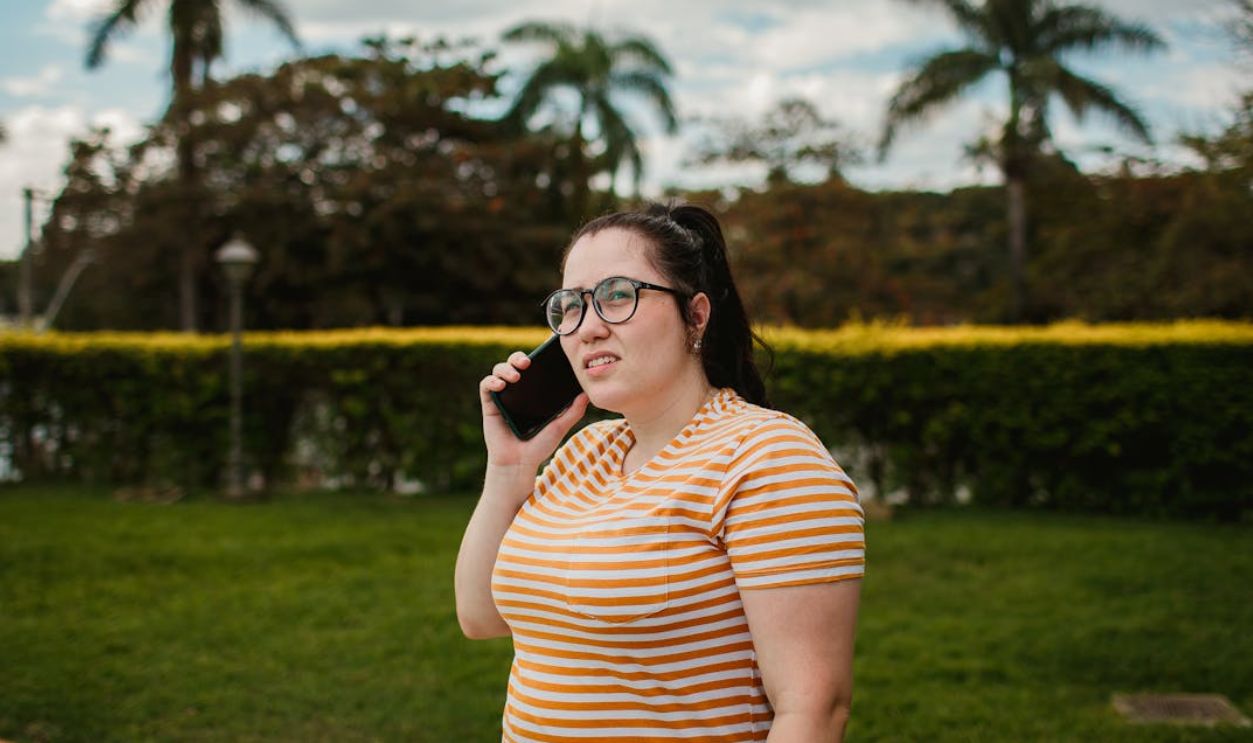 Woman in striped shirt talks on phone outside in a lush green park