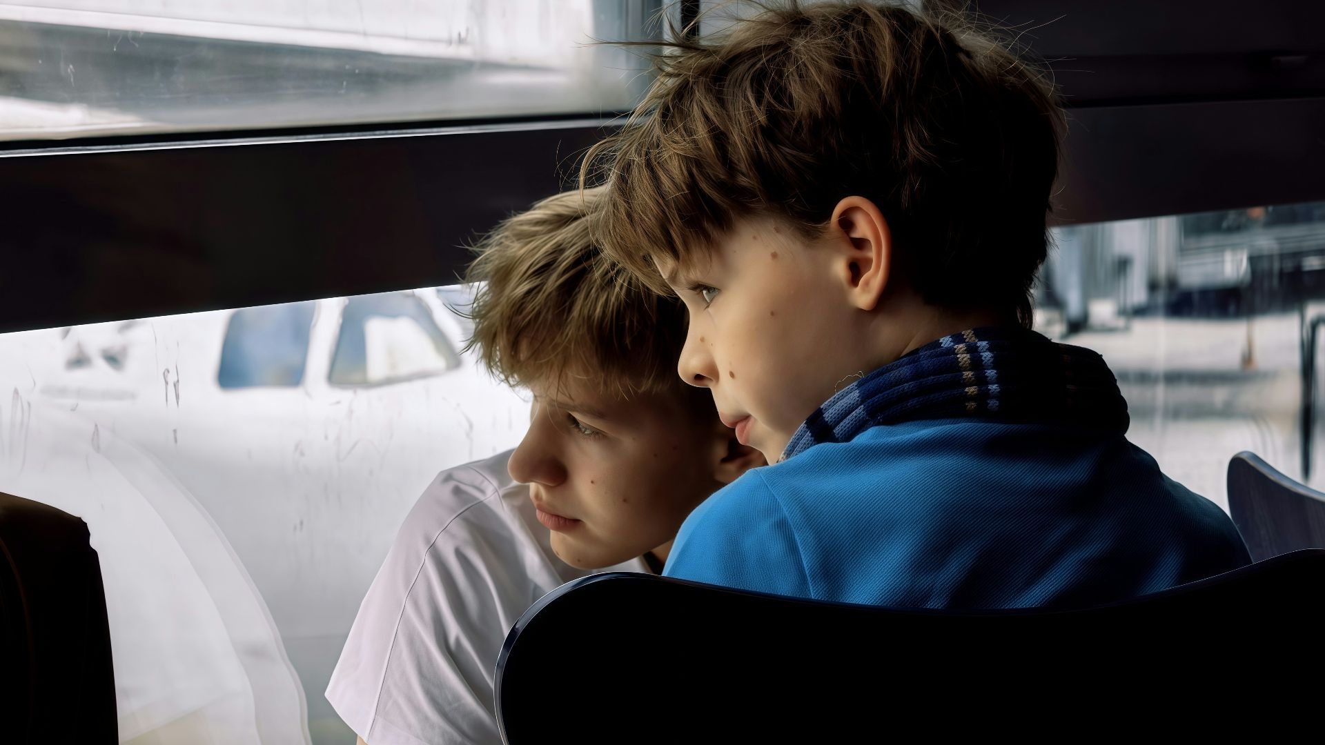 Two boys look out airport window at airplane.