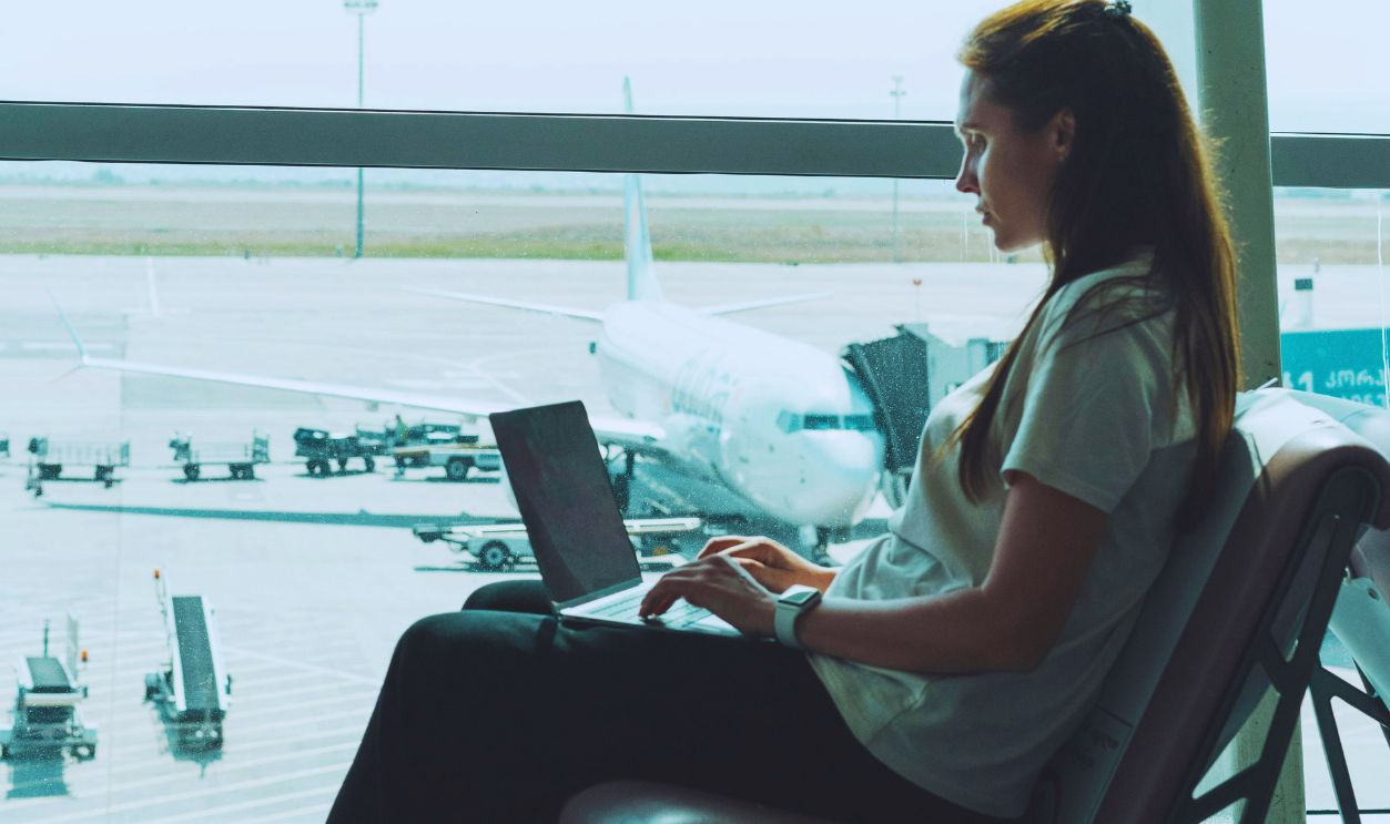 Side view of woman using laptop at airport, airplane visible outside through large glass windows