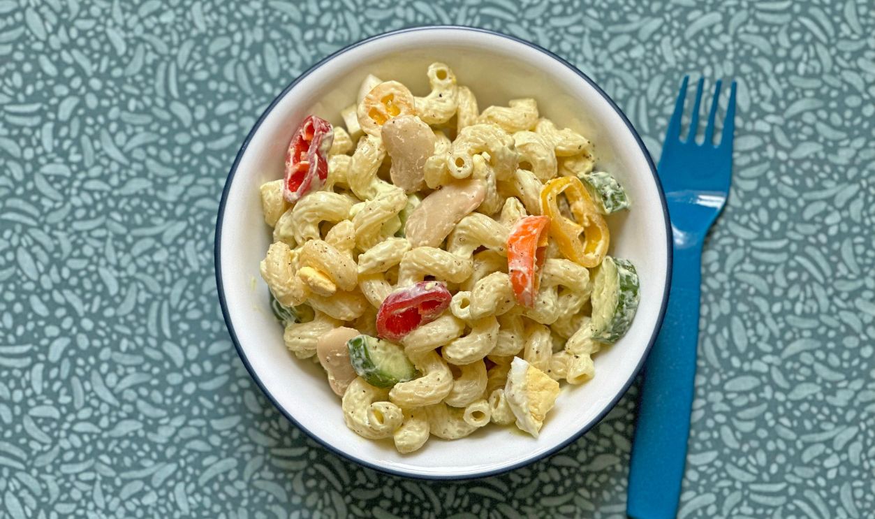 Getty Images - 2260533179 - A cold protein pasta salad with vegetables and creamy avocado dressing, served in a bowl with a blue fork, on a patterned tablecloth.