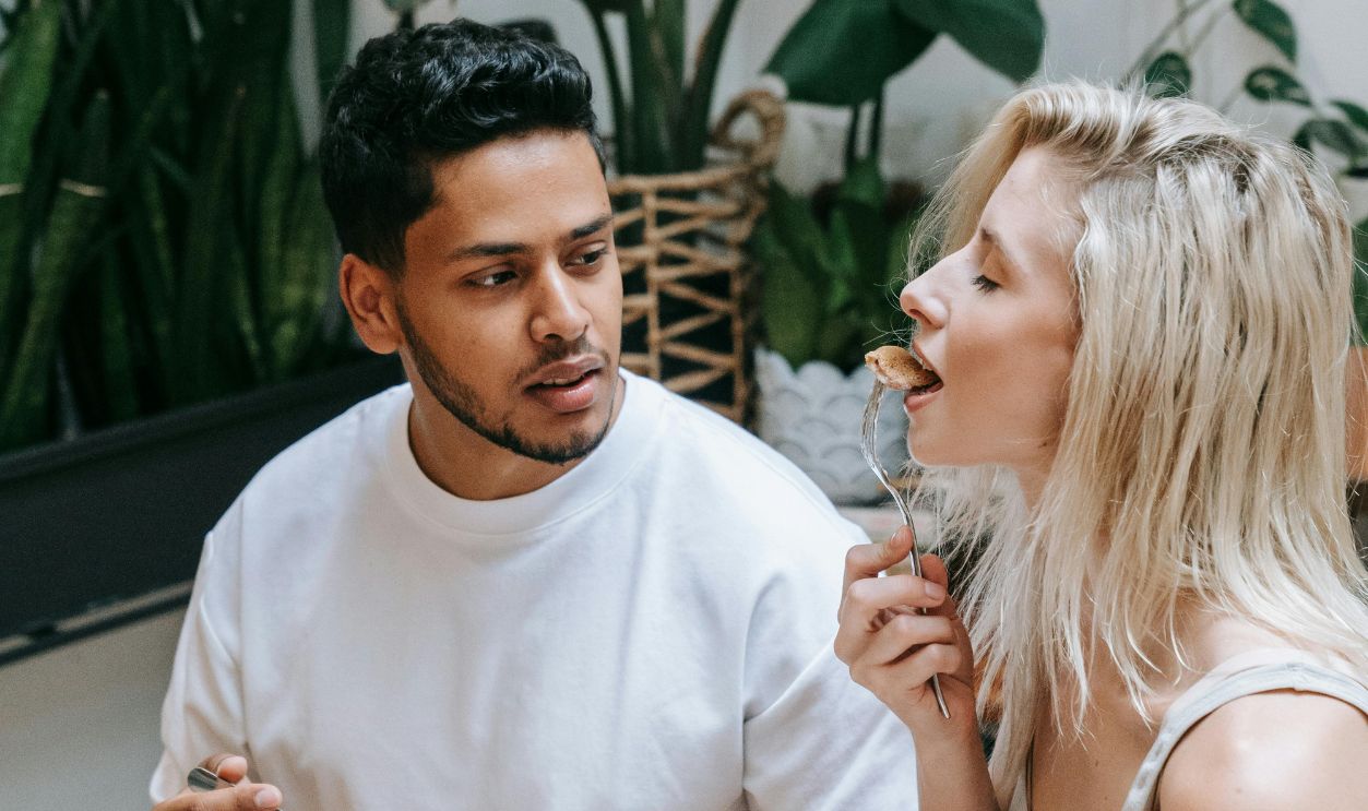 An interracial couple happily sharing a breakfast meal indoors with lush plants.