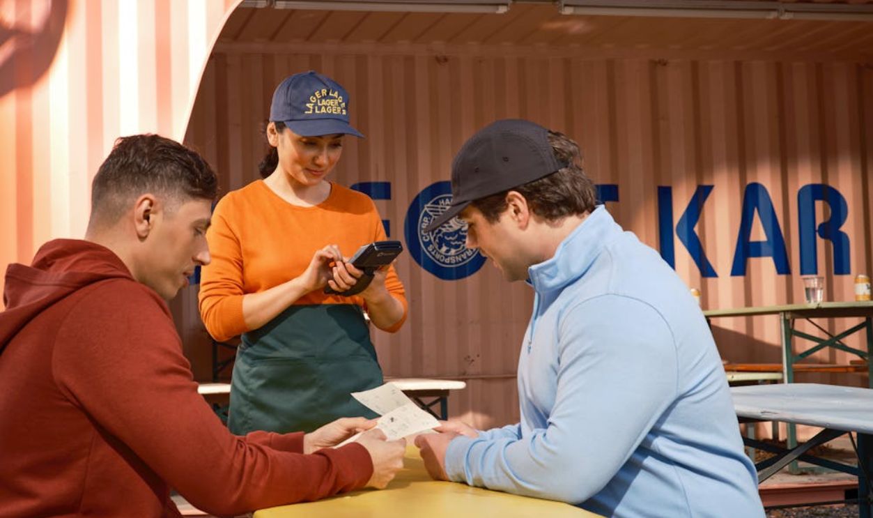 A restaurant staff member takes orders using a handheld device in an outdoor cafe setting.