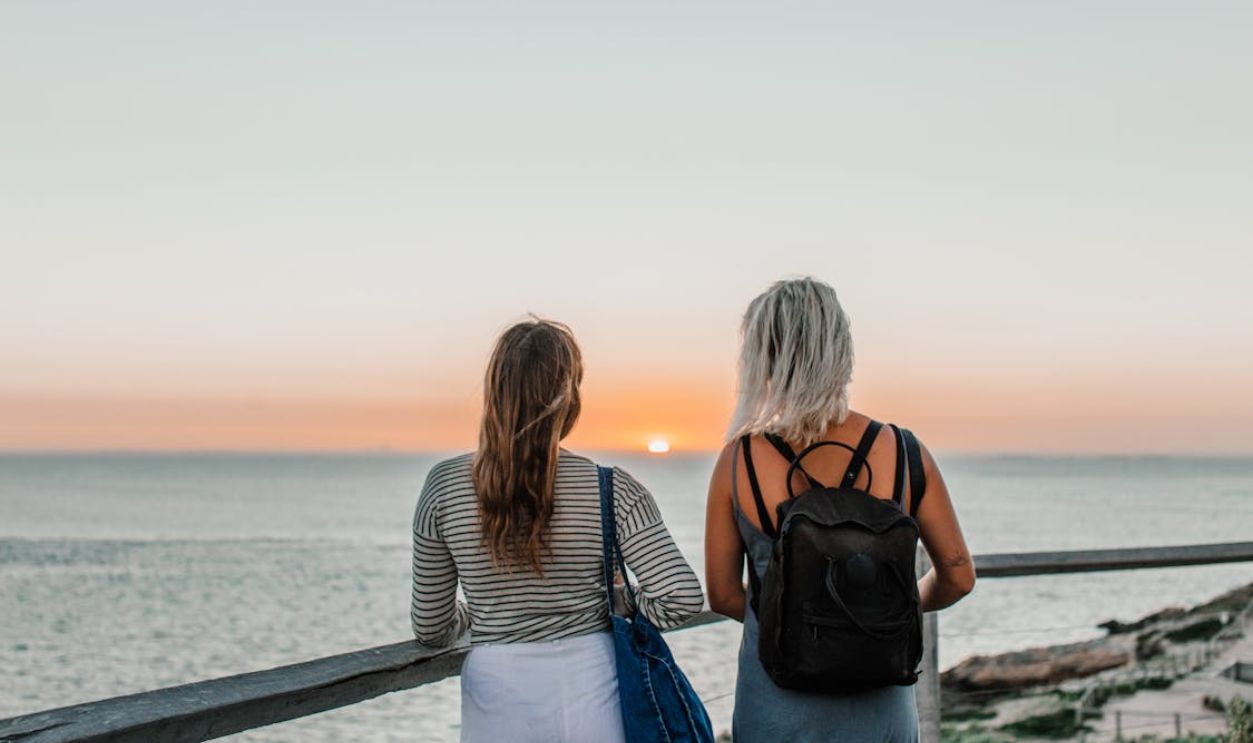 Two women with backpacks watching a serene ocean sunset from a wooden deck.