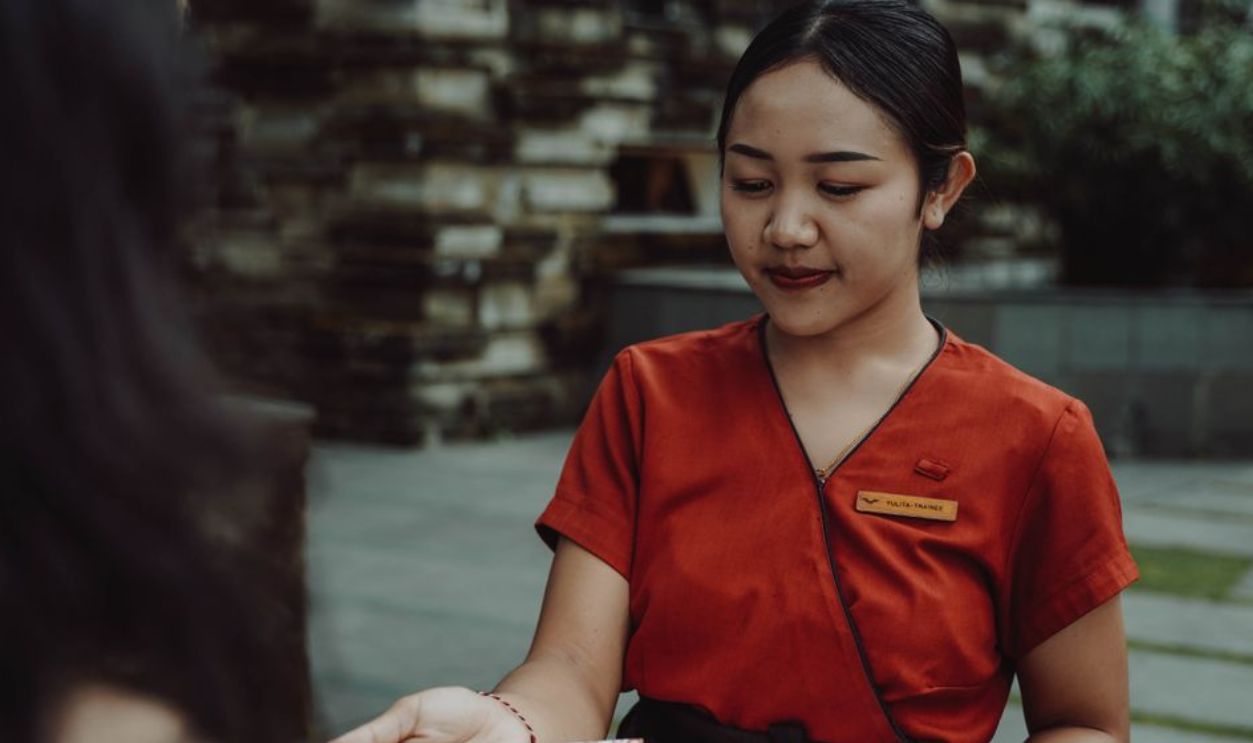 A waitress in a red uniform handing a menu to a guest at an outdoor restaurant