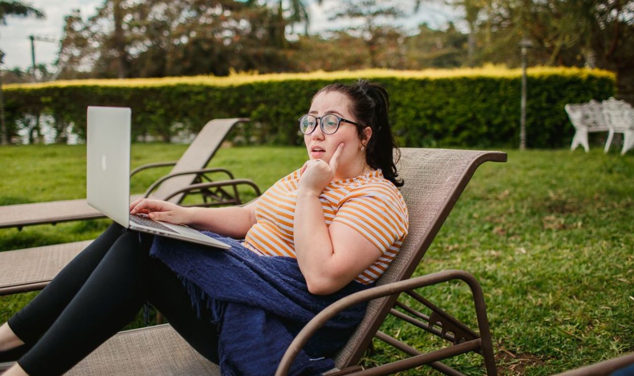 Woman relaxing on lounge chair with a laptop in a lush green garden, enjoying a sunny day