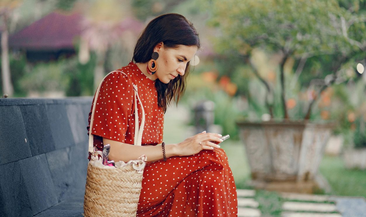 Woman in a red polka dot dress using a smartphone while sitting on a bench outdoors.