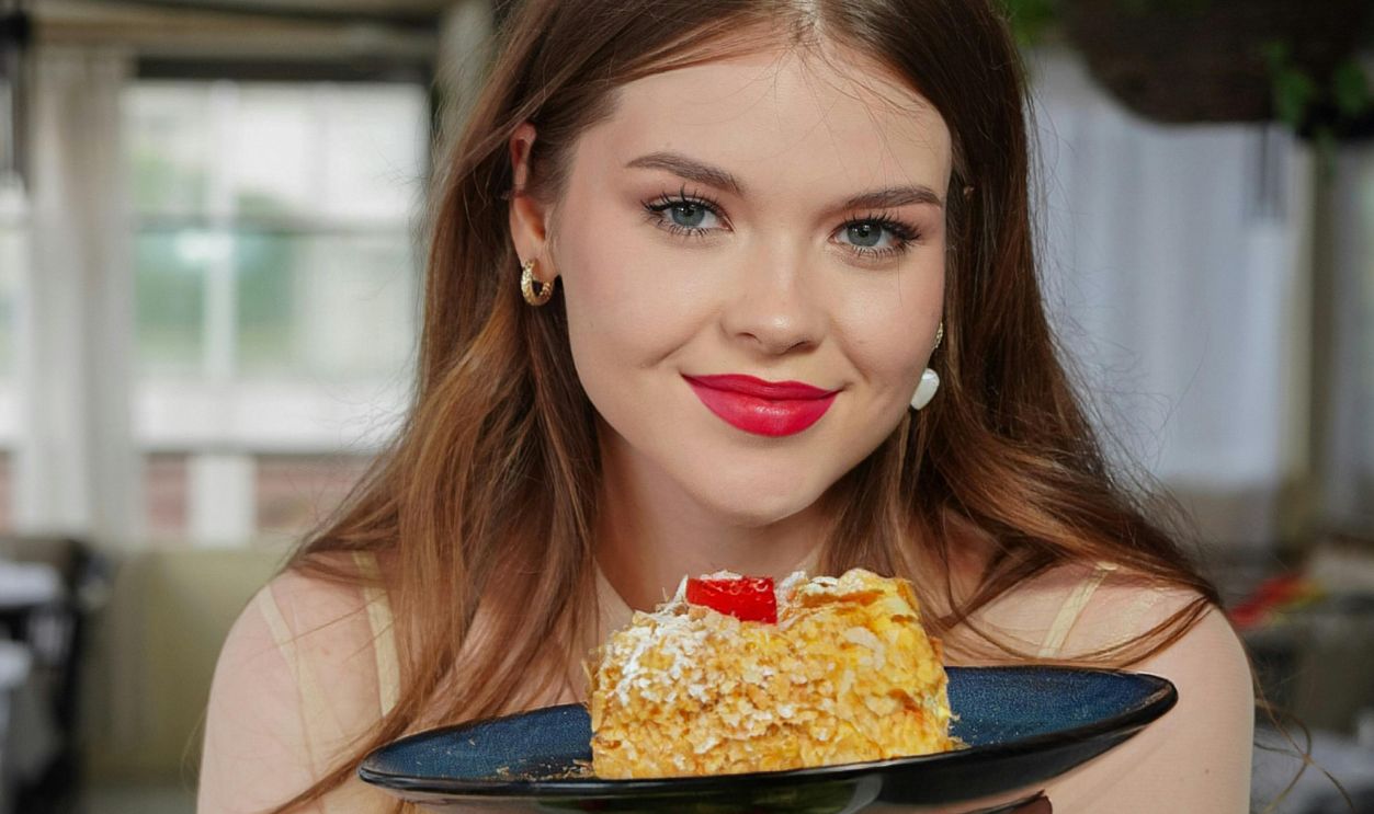 Smiling woman with long brown hair holding a slice of cake and a bowl of salad indoors.
