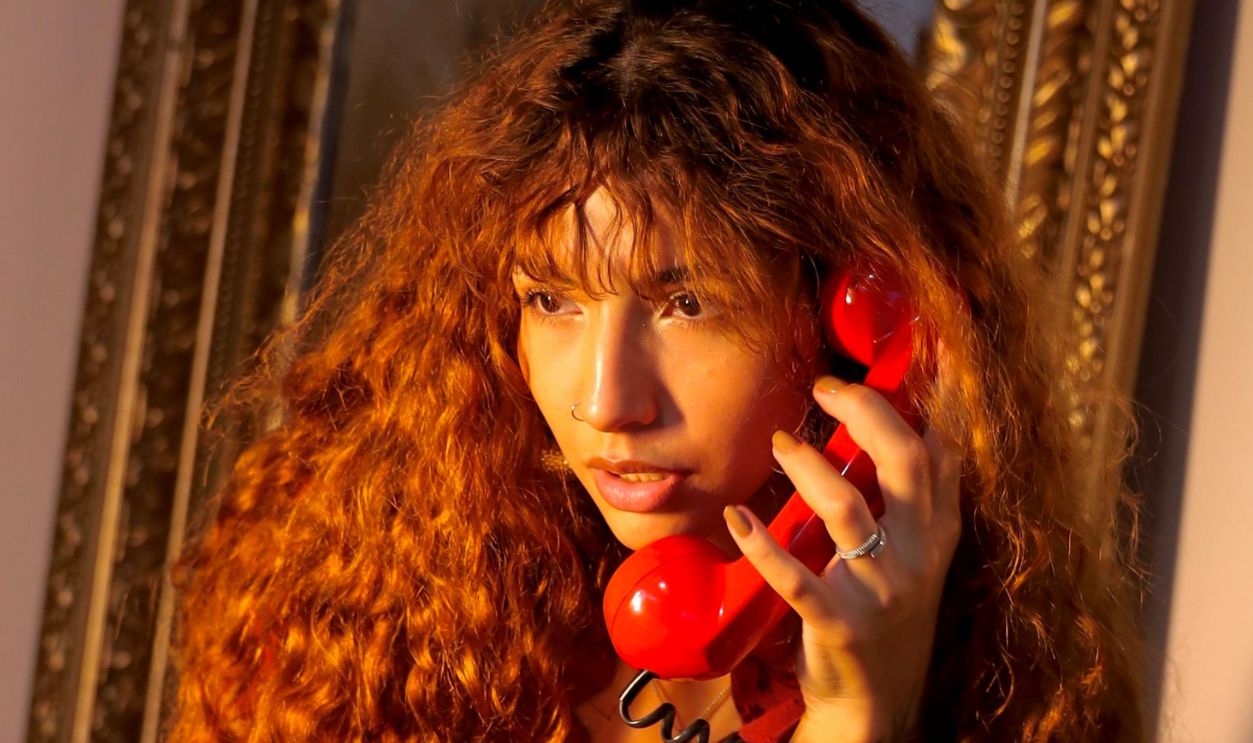 Portrait of a woman with red curly hair sitting indoors, holding a retro red phone