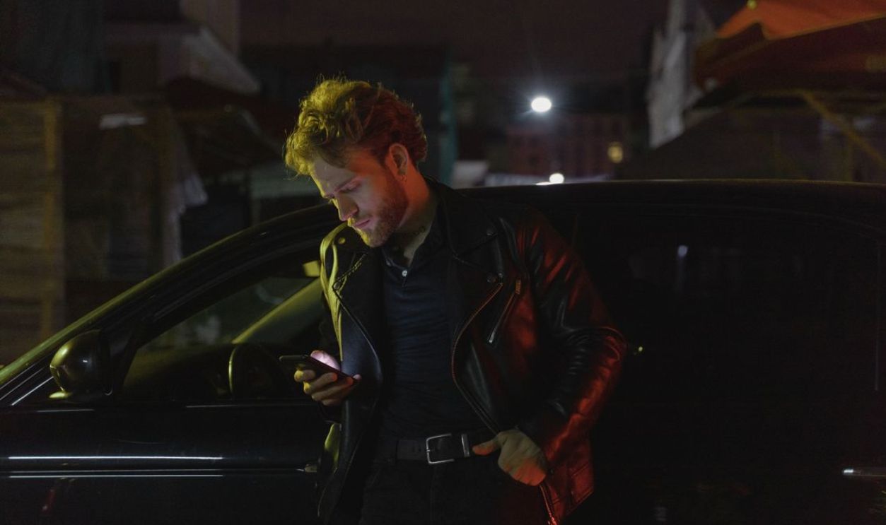 Caucasian man stands by a car at night, using his phone. Retro 80s style with a moody atmosphere