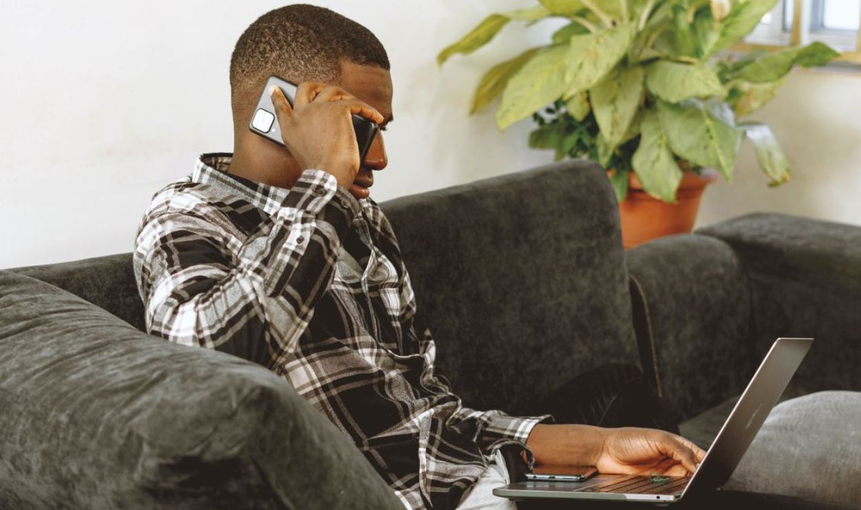 Young man multitasking with phone and laptop on a sofa, working remotely from home