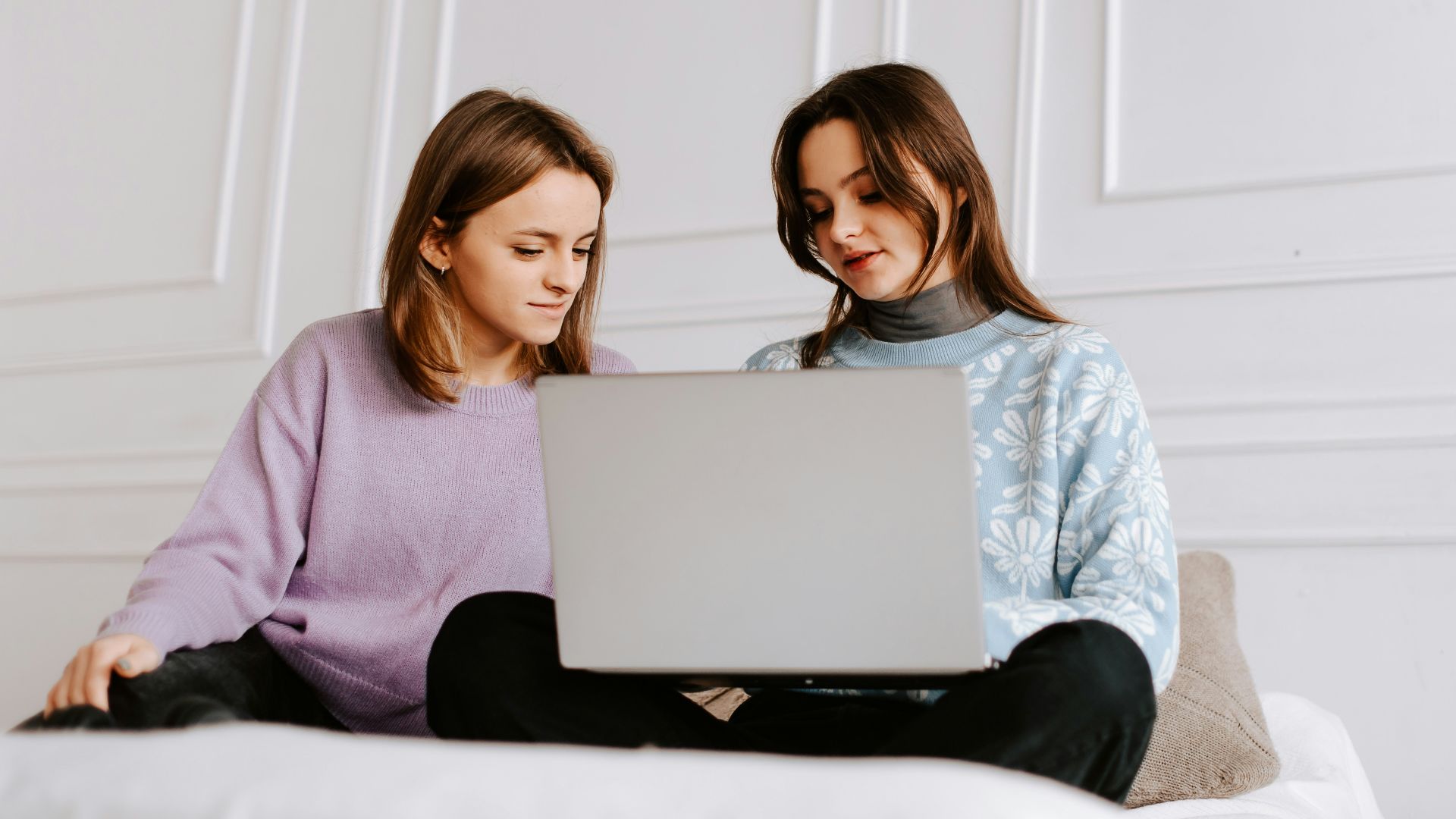 two women sitting on a bed looking at a laptop