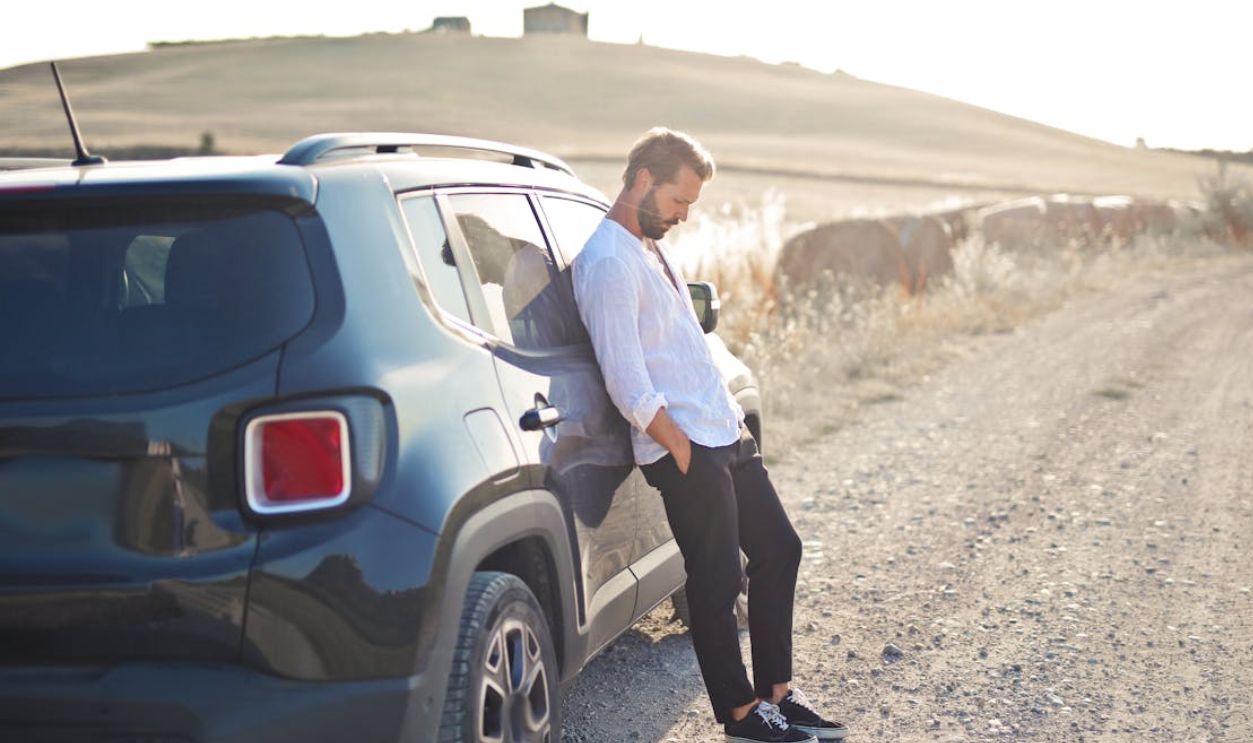 A man in a white shirt leans against a car on a sunny rural dirt road. Simplicity and solitude.