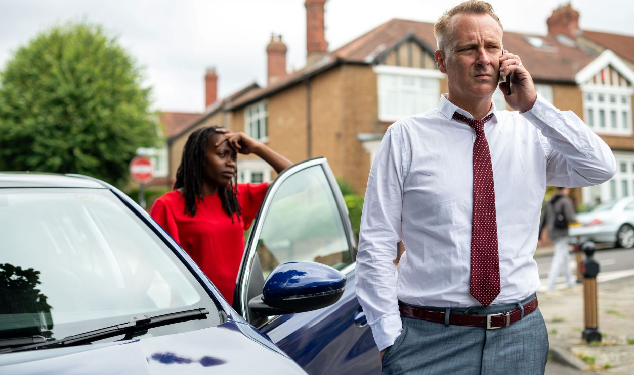 A man in a white shirt and red tie talks on the phone beside a car. A woman in a red shirt stands nearby, looking concerned in a suburban neighborhood.