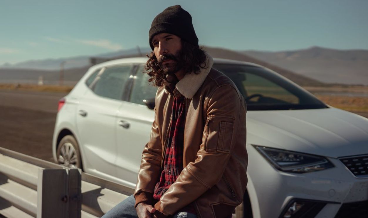 Man in a beanie sitting by a car on an open road with mountains in the background