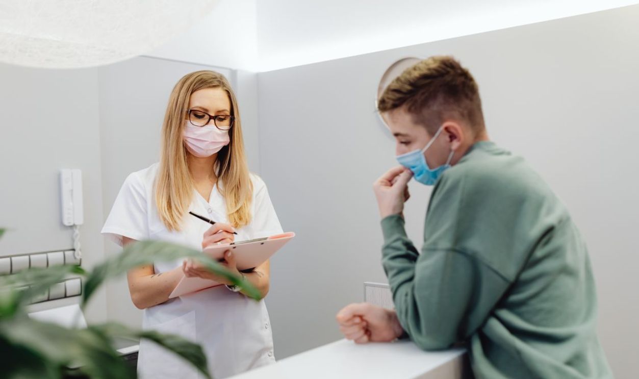 A healthcare worker consults a patient at a clinic reception, both wearing face masks.