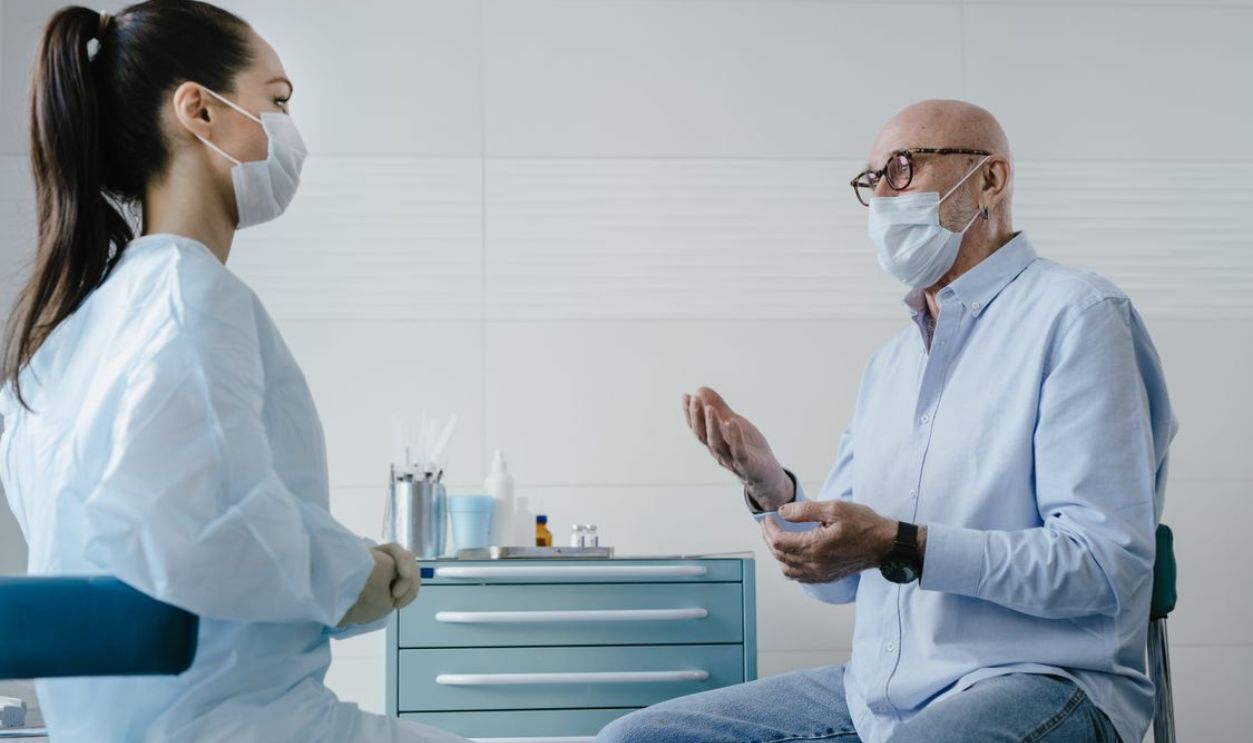 A senior adult patient discusses health concerns with a masked medical professional in a clinic setting.