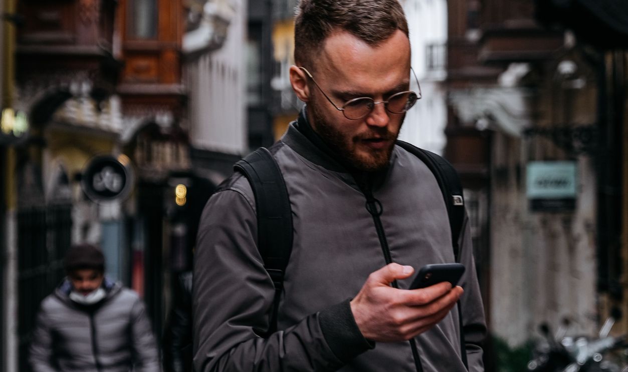 A man with a backpack attentively checks his smartphone while walking on a cobblestone street