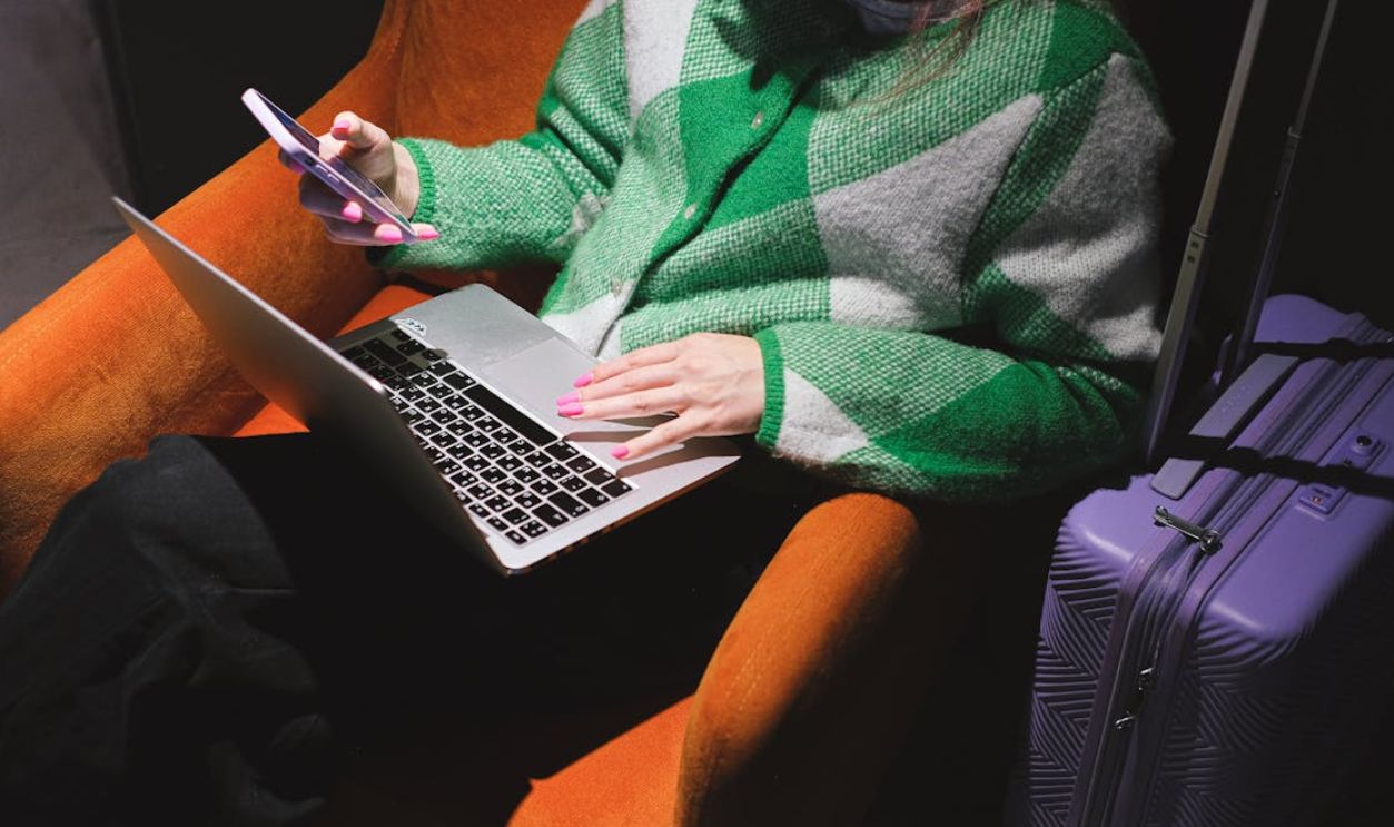 Woman in a knitted sweater working remotely on laptop with a smartphone, sitting in an armchair
