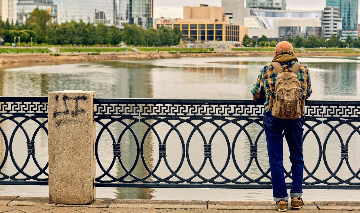 A man stands by a metal fence, overlooking a river in an urban setting with modern buildings