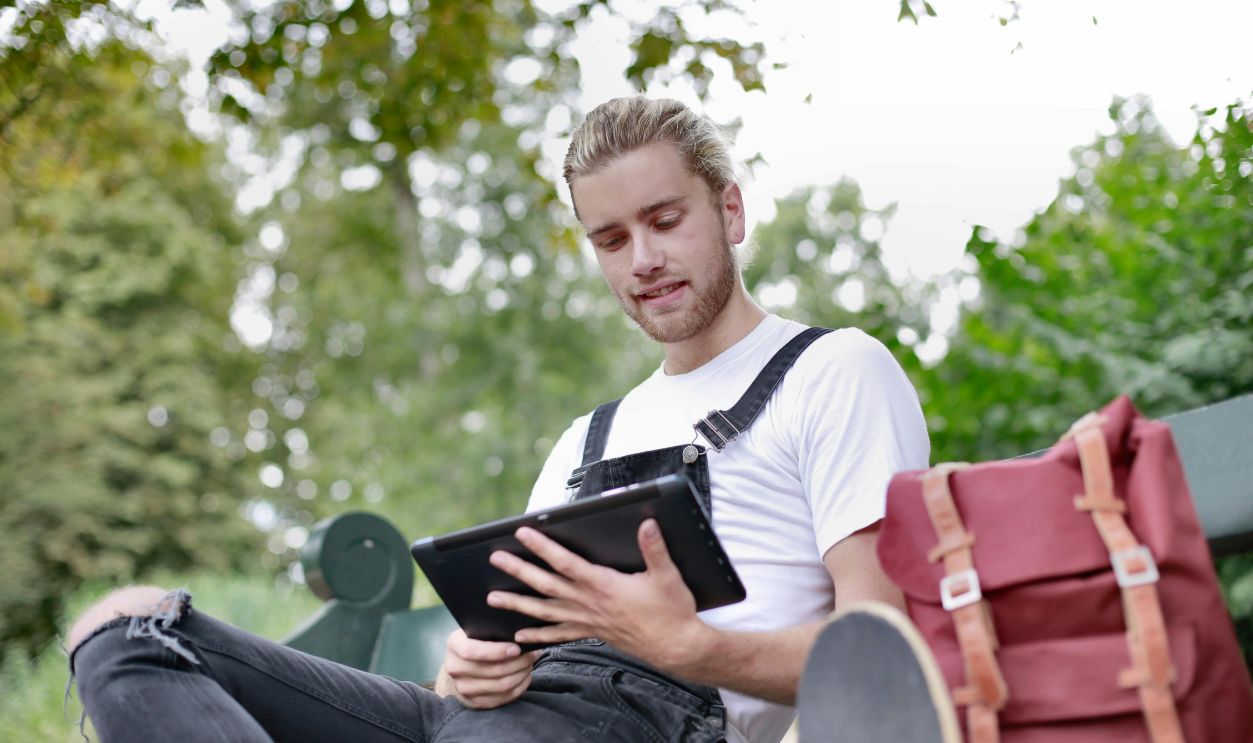 Young man sitting on bench in park using tablet with backpack nearby, enjoying leisure time outdoors