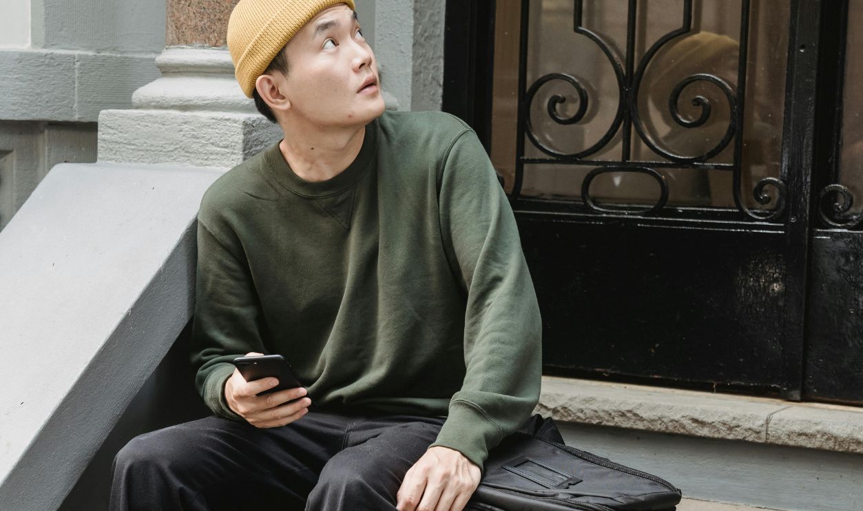 Fashionable young man in beanie sitting on steps outdoors, looking up, holding a smartphone