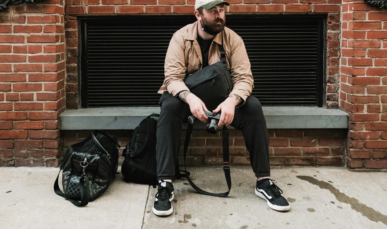 Man with bag and camera sits against a brick wall in an urban setting
