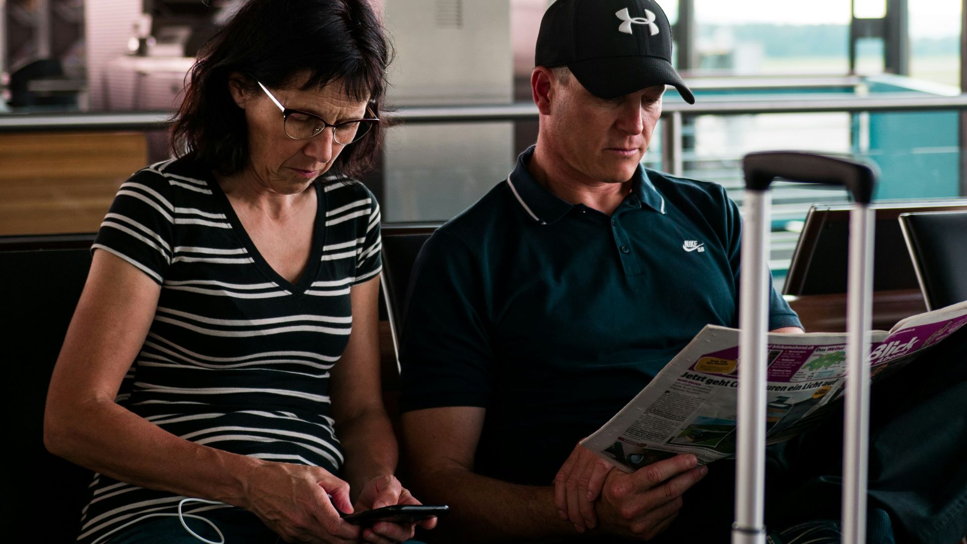 woman fiddling on smartphone beside man with cap reading newspaper