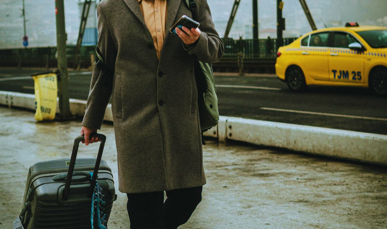 A man walking outdoors with a suitcase and smartphone near yellow taxis on a city street