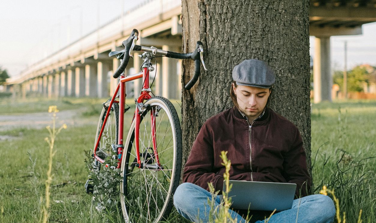 Caucasian man sitting under a tree with a laptop and road bike beside an overpass.