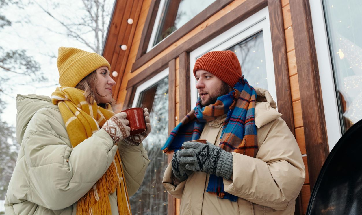 A couple in winter attire enjoys hot drinks outside a cozy cabin. Perfect winter bonding scene