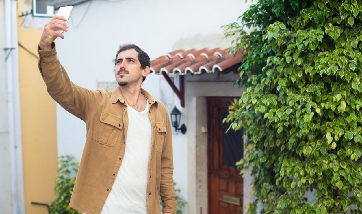 A man in a brown jacket takes a selfie outdoors, capturing travel memories.